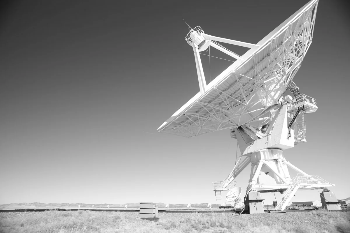 Grande antenna parabolica bianca puntata verso un cielo sereno, con altre antenne identiche allineate sull’orizzonte, fotografia in bianco e nero