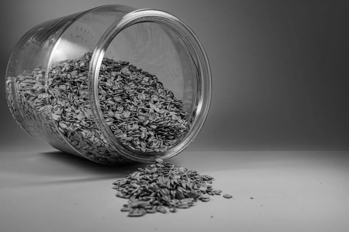 A glass jar tipped on its side, with rolled oat flakes spilling out and scattered across the surface, black-and-white photograph