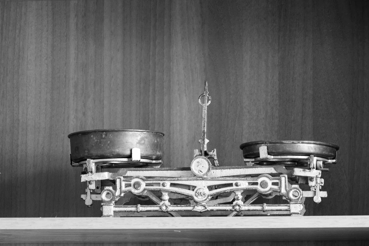 A brass two-pan balance scale on a wooden shelf, its cast-iron frame stamped with the load capacity, black-and-white photograph