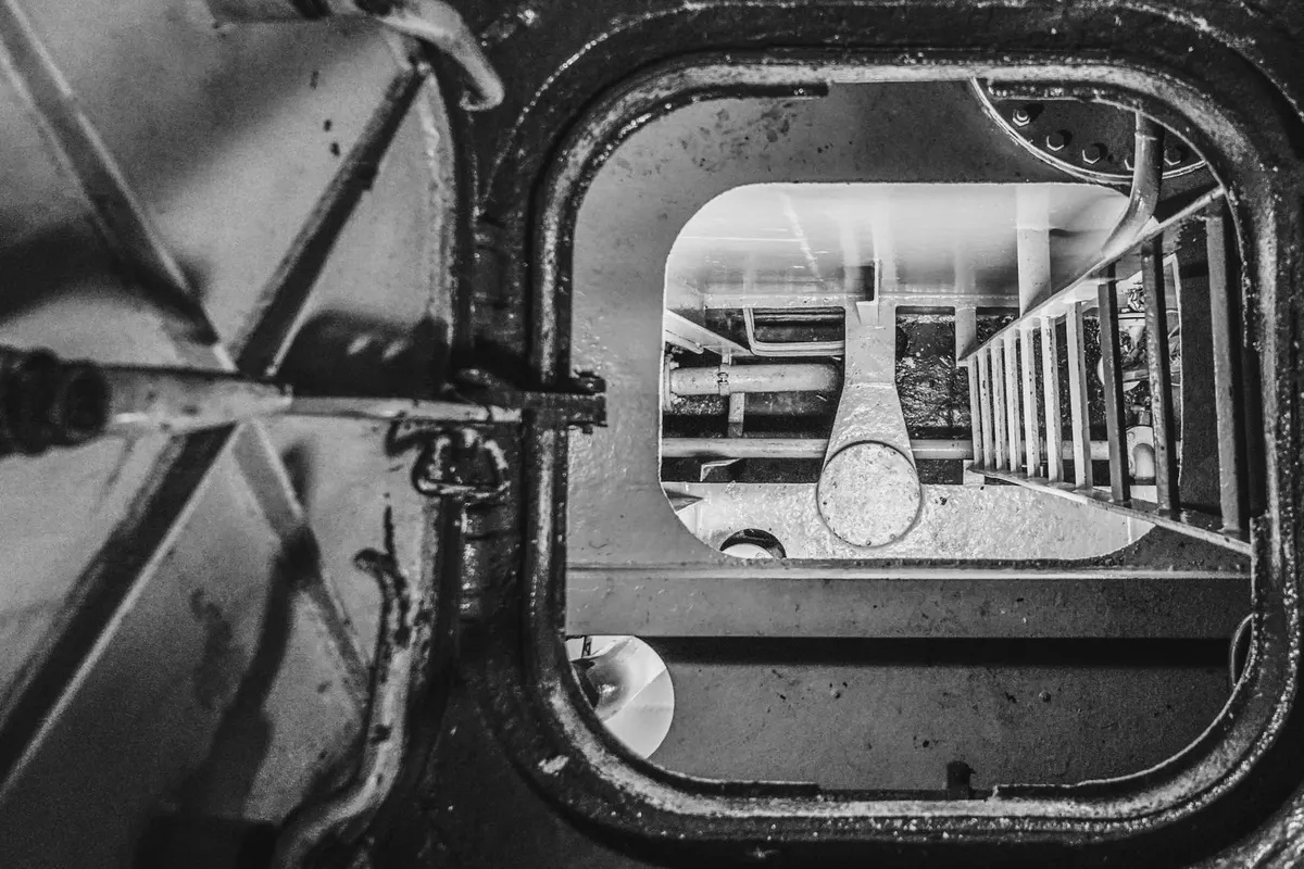 A ship’s metal hatch with its cover swung wide open, revealing pipework and a ladder in the hold below, black and white photograph