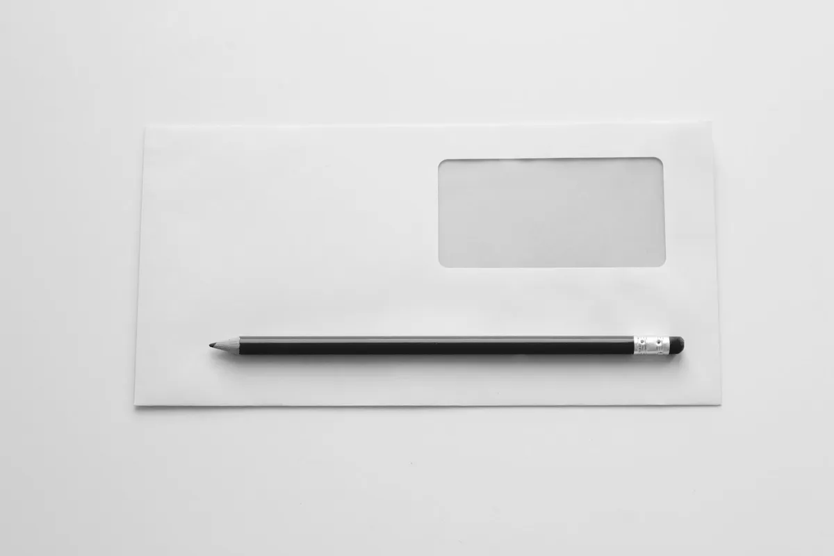 A closed white envelope with an empty transparent window and a black pencil resting on top, in black and white