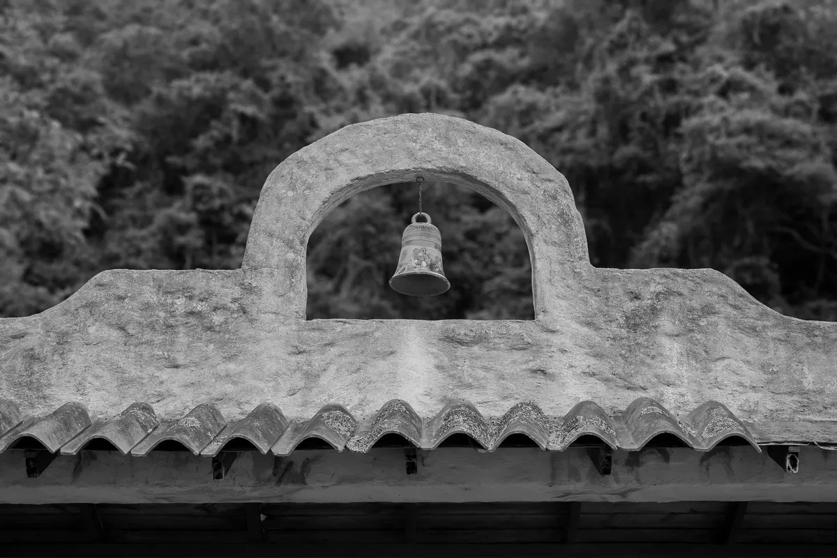 Piccola campana appesa a una vela campanaria in muratura sopra un tetto di coppi, fotografia in bianco e nero; nessuna scritta, nessun marchio, nessuna persona