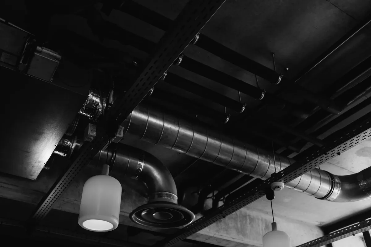 Air ducts, pipework and cable trays beneath a building ceiling, with a smoke detector, black-and-white photograph