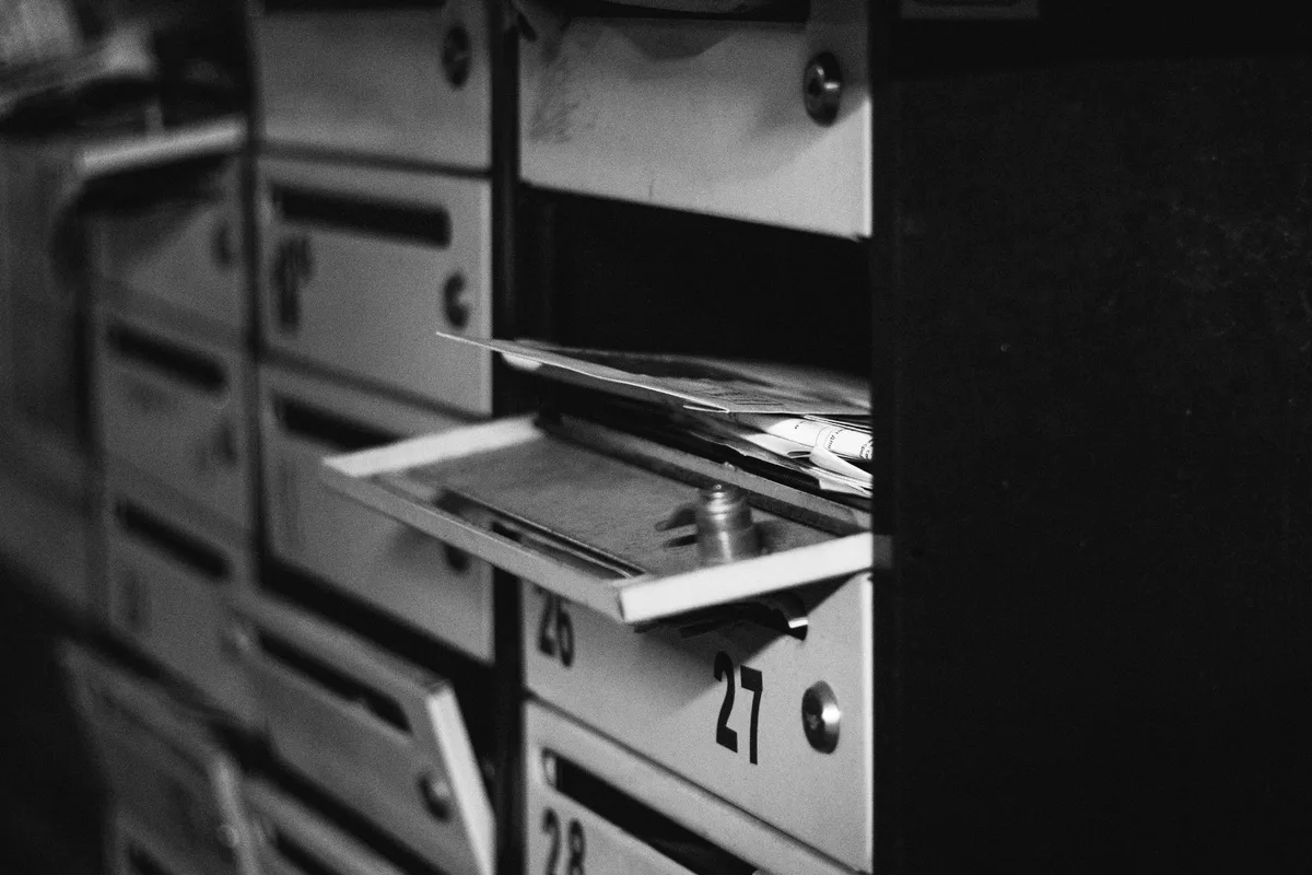 Fila di caselle postali metalliche numerate in sequenza, una aperta con dentro la posta, in bianco e nero