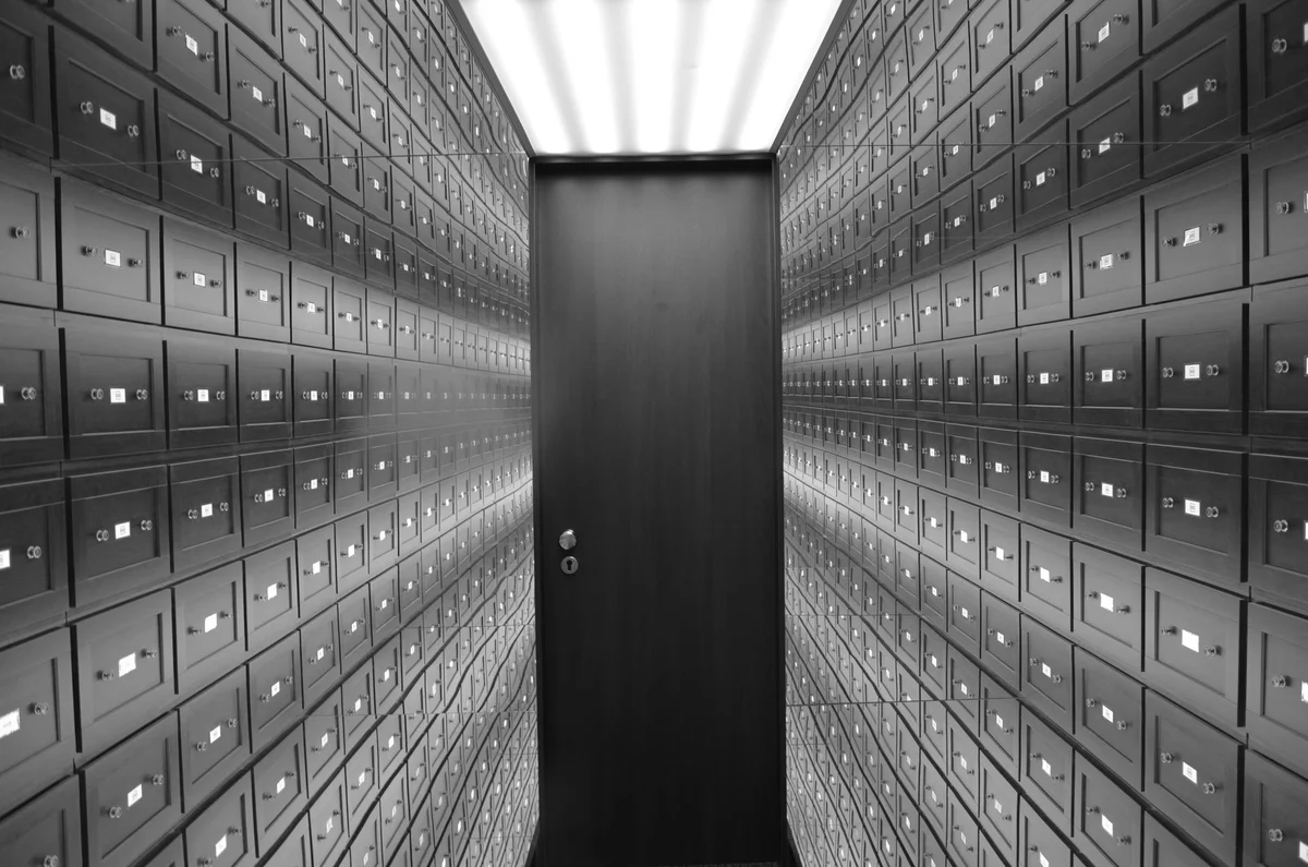 Corridoio di una sala caveau con file di piccole cassette di sicurezza in legno scuro, ciascuna con targhetta e serratura, che convergono verso un’unica porta blindata in fondo, fotografia in bianco e nero