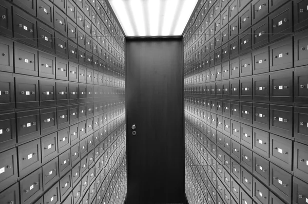 Corridoio di una sala caveau con file di piccole cassette di sicurezza in legno scuro, ciascuna con targhetta e serratura, che convergono verso un’unica porta blindata in fondo, fotografia in bianco e nero