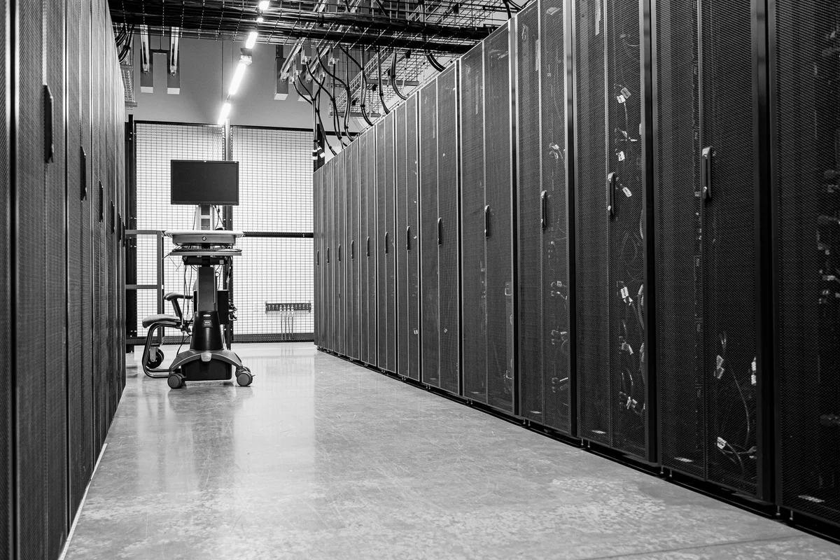 Corridor of a data centre with rows of closed server cabinets, cables suspended from the ceiling, and a mobile workstation with a blank monitor, in black and white