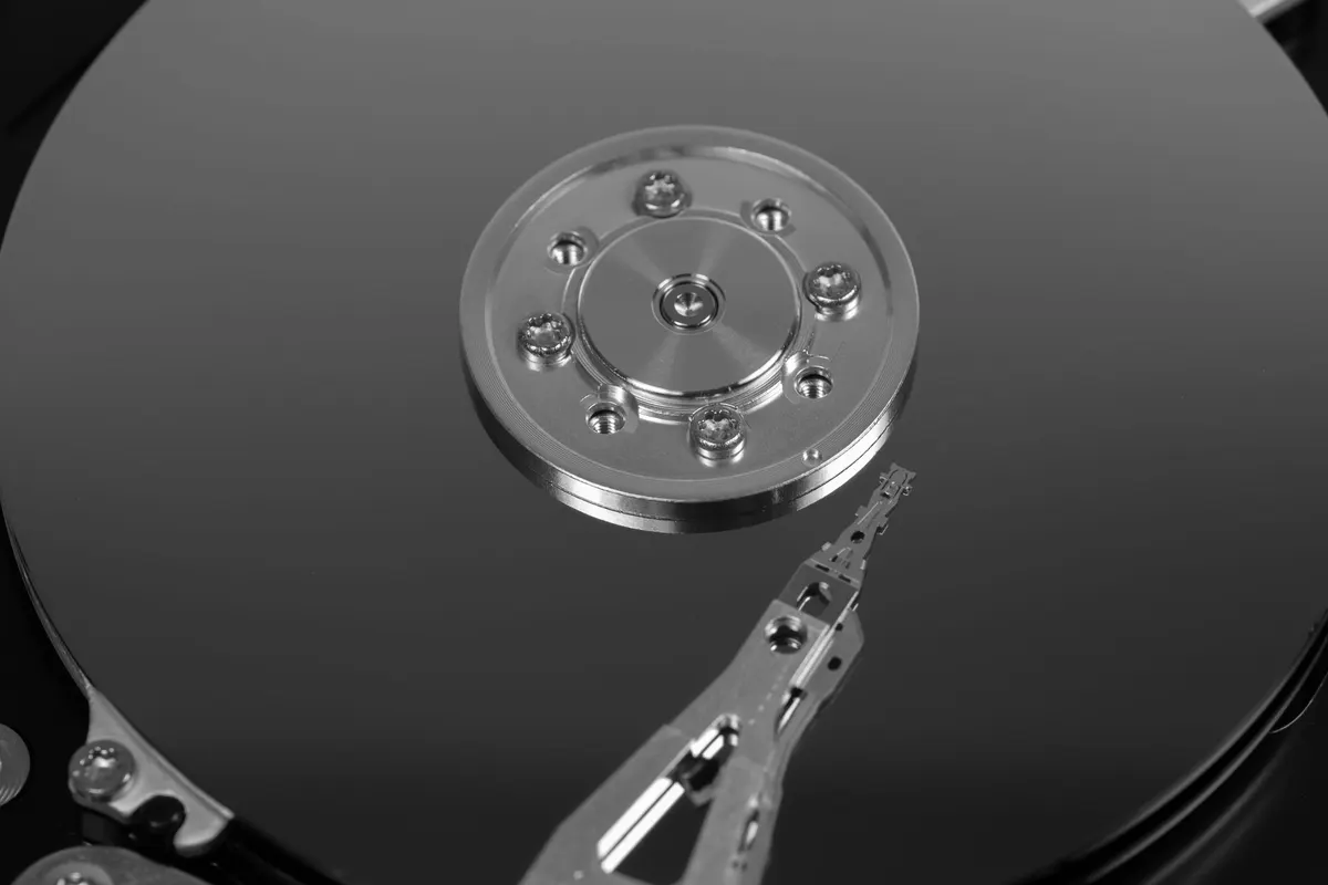 Primo piano della testina di lettura sospesa sopra il piatto metallico riflettente di un disco rigido aperto, con le viti del fermo centrale disposte in cerchio attorno al mozzo, fotografia in bianco e nero