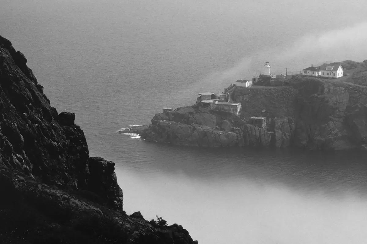 A rocky cliff overlooking a sea strait in fog, with a lighthouse and a few white buildings on a headland, black and white photograph