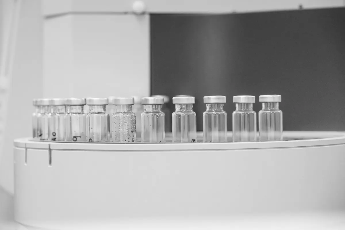 Fila di fiale di vetro con tappo metallico allineate su un vassoio circolare di uno strumento da laboratorio, sullo sfondo una parete scura sfocata, fotografia in bianco e nero