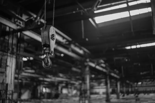 Gancio di paranco appeso a una fune d’acciaio dentro un capannone industriale in penombra, senza alcun carico agganciato, fotografia in bianco e nero, nessuna persona