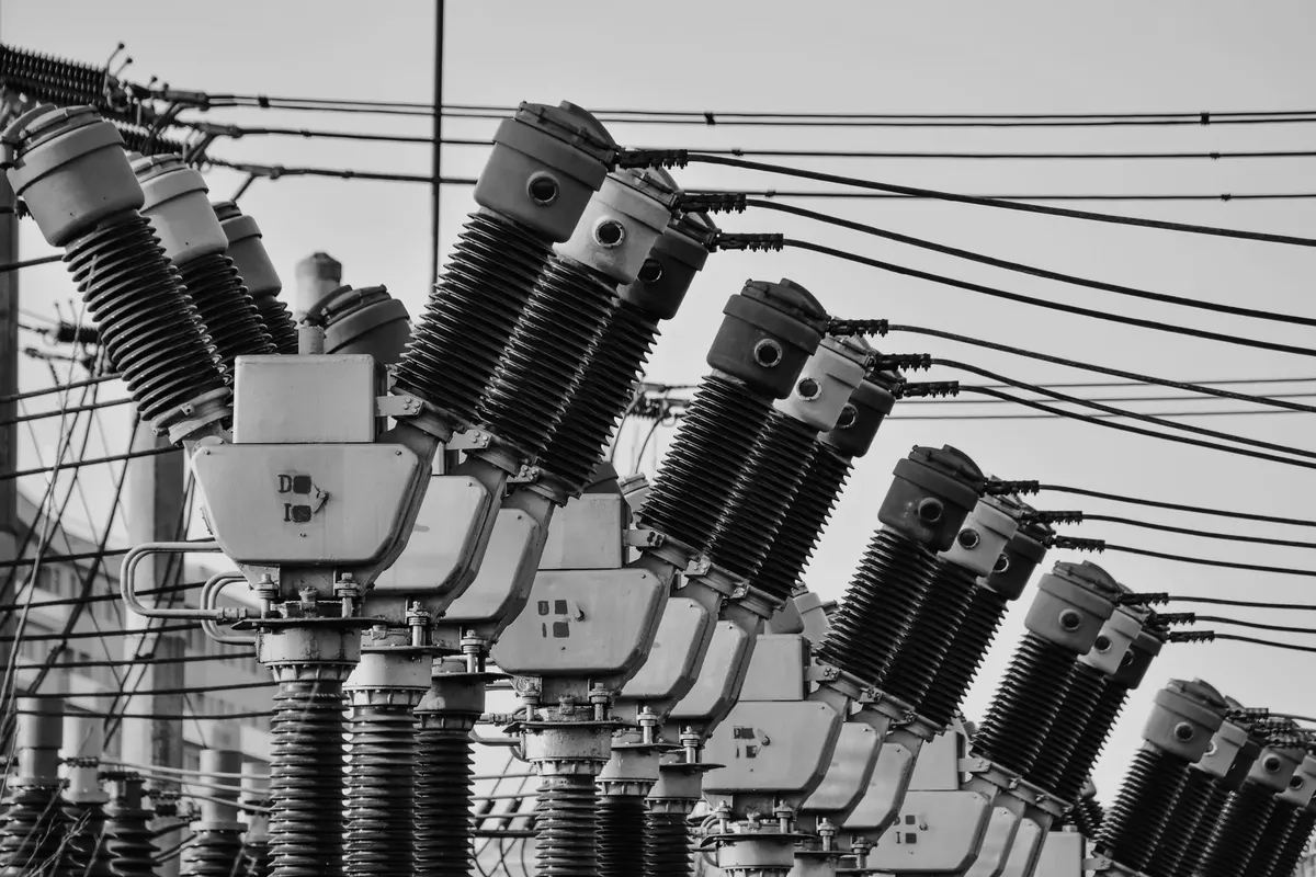Una fila di sezionatori e isolatori ad alta tensione in una sottostazione elettrica, cavi tesi contro un cielo uniforme, fotografia in bianco e nero