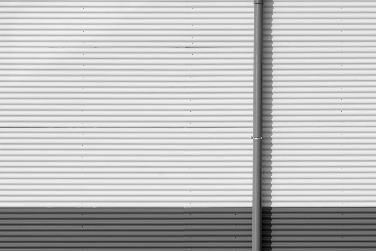 Corrugated metal wall of an industrial building, photographed frontally in black and white, with a vertical downpipe