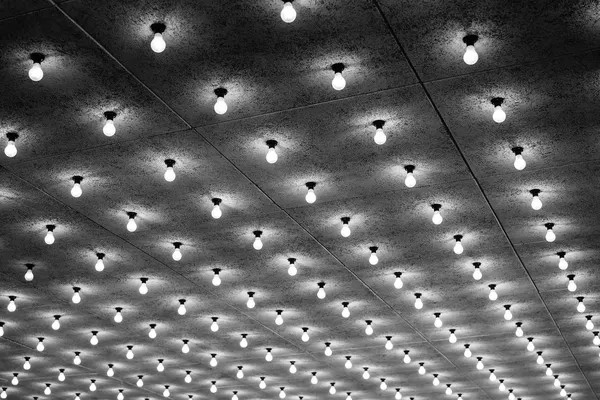 Griglia fitta di lampadine nude e accese, vista dal basso su un soffitto a pannelli, fotografia in bianco e nero