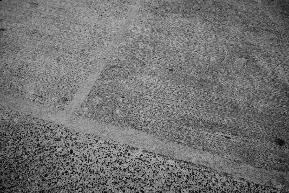 Due linee chiare tracciate su un pavimento industriale in cemento, che separano una superficie liscia da una a grana grossa, fotografia in bianco e nero