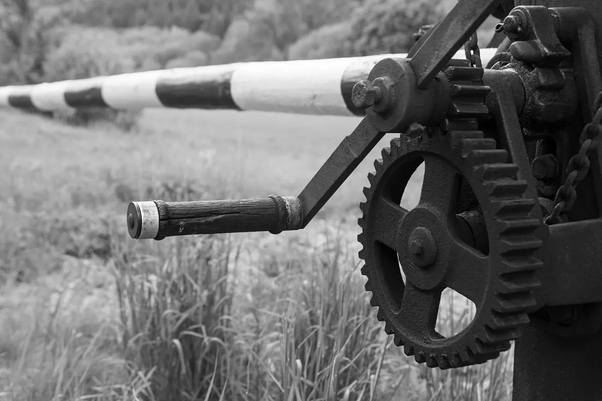 Crank handle and gear of a hand-operated striped boom barrier, with the arm lowered in the background, black and white photograph