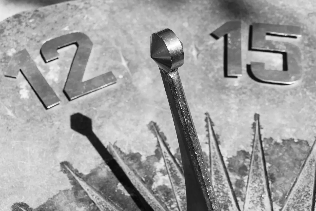 Primo piano dello gnomone metallico di una meridiana in pietra, con la sua ombra proiettata accanto alle cifre metalliche applicate "12" e "15", fotografia in bianco e nero