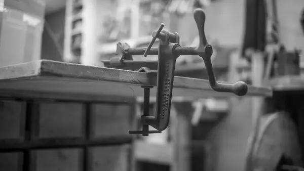 Morsetto a vite in ghisa serrato sul bordo di un banco da lavoro in legno, officina sullo sfondo, fotografia in bianco e nero