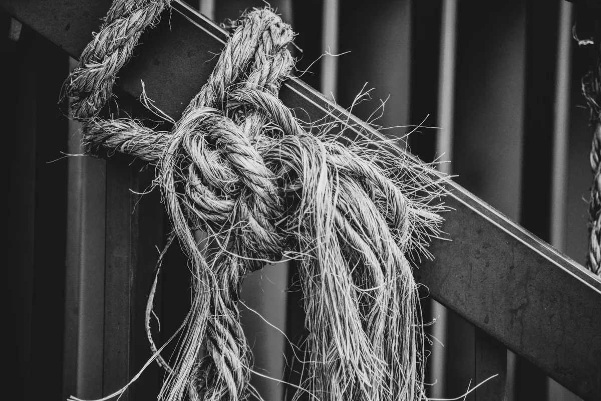 Worn, frayed rope knot tied to a metal beam, in black and white