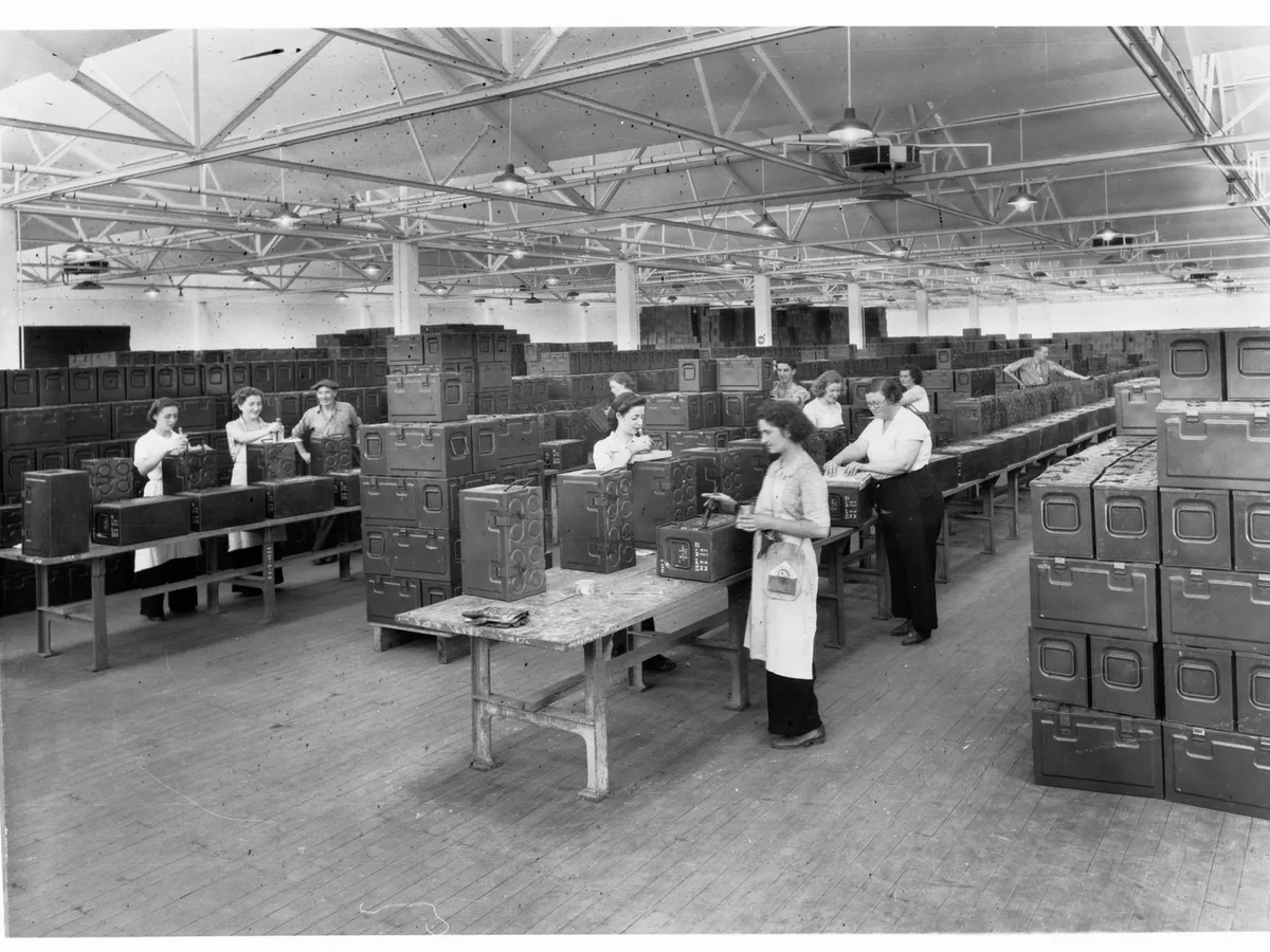 Period interior of a munitions factory: workbenches and long rows of stacked metal crates
