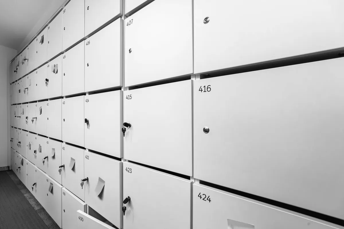 Parete di armadietti numerati lungo un corridoio, con alcune chiavi lasciate nelle serrature e foglietti adesivi sugli sportelli, in bianco e nero