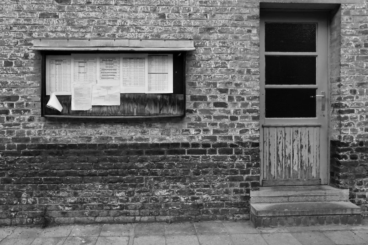 A public notice board in a wooden glass case on a brick wall, sheets pinned behind the glass, a closed door beside it, in black and white