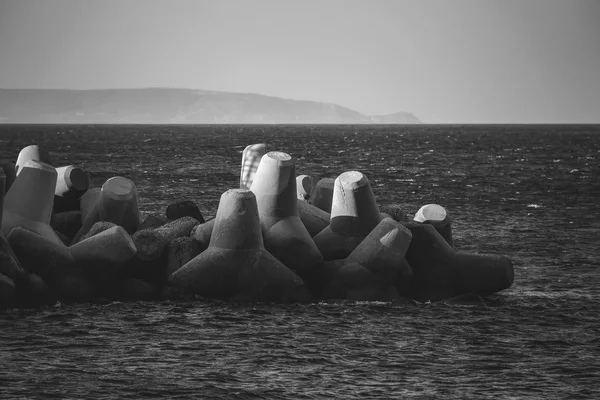 Breakwater of stacked concrete blocks facing the open sea, with a headland on the horizon, in black and white