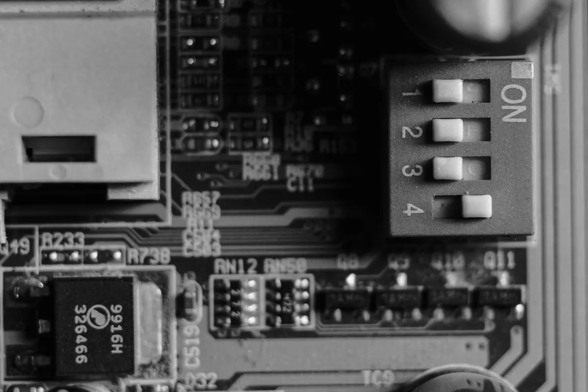 A DIP switch block on a circuit board, three levers in one position and the fourth in the other, in black and white