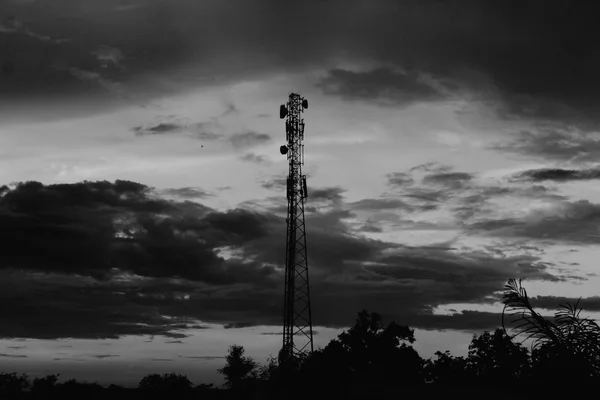 The silhouette of a telecommunications mast against a cloudy sky at dusk, in black and white