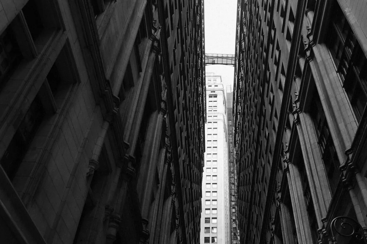 A sliver of sky between two adjoining tower blocks, an elevated walkway crossing it high up, black-and-white photograph