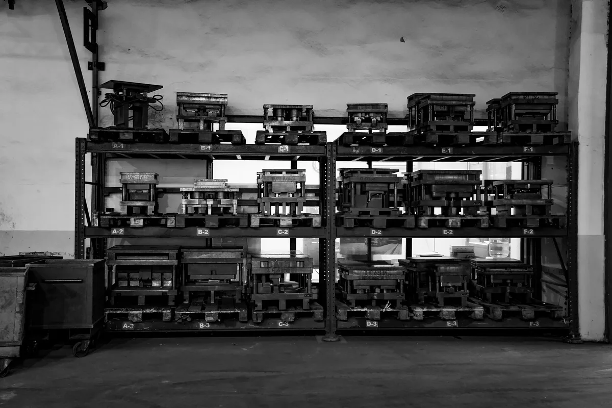 Scaffalatura industriale in officina con stampi metallici da pressa disposti su ripiani contrassegnati da lettere e numeri, in bianco e nero