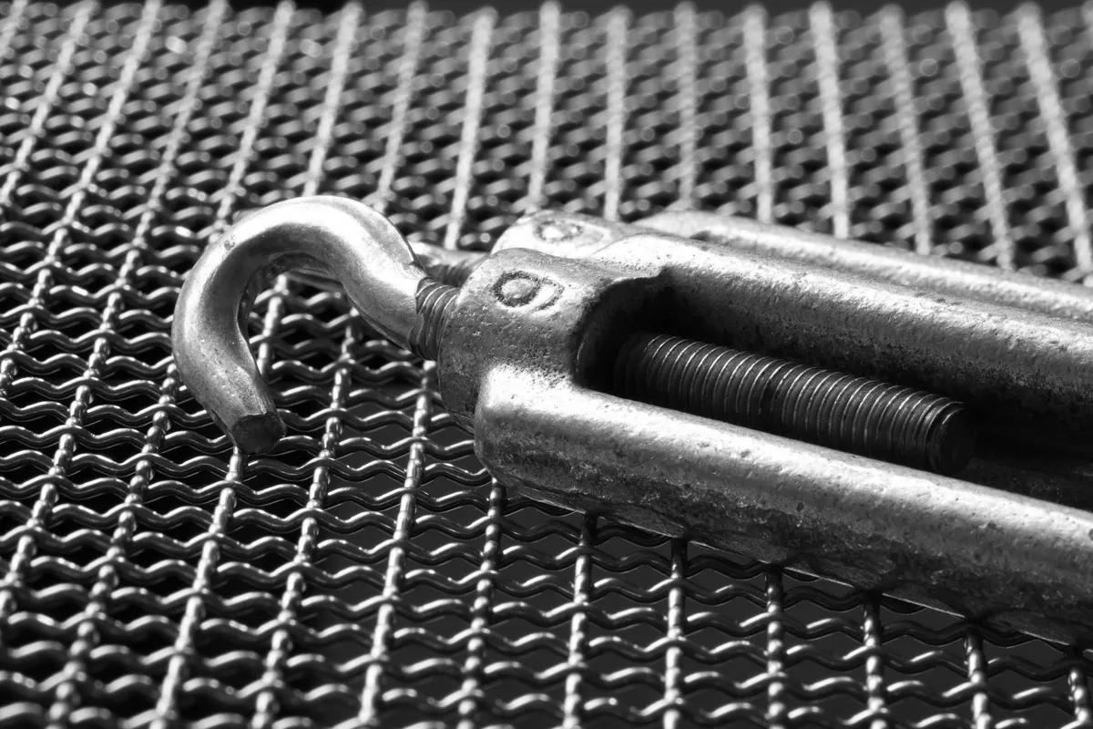 Close-up of a steel turnbuckle with a hook resting on wire mesh, black and white photograph