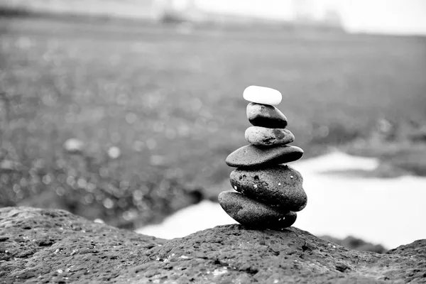 Pila di sassi in equilibrio su una roccia più ampia, sullo sfondo una spiaggia sfocata, fotografia in bianco e nero senza persone né scritte