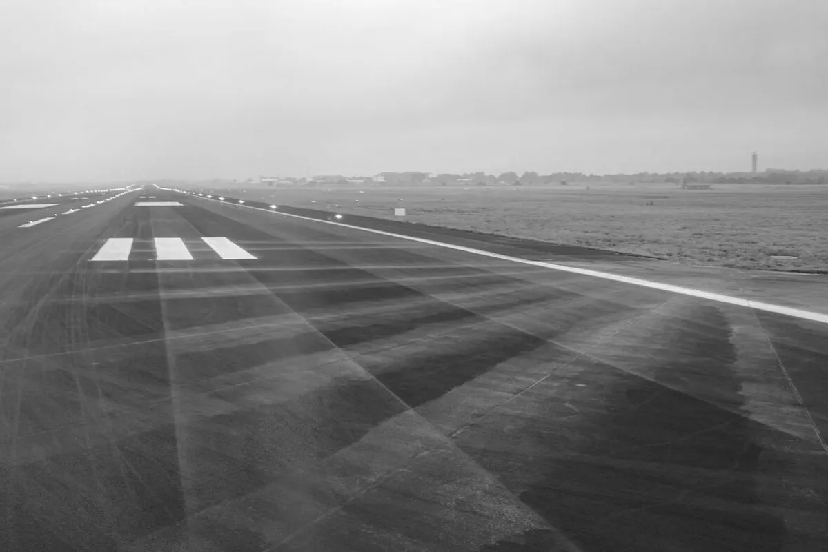 The threshold of an airport runway with painted markings on the tarmac, countryside and overcast sky in the background, black and white photograph