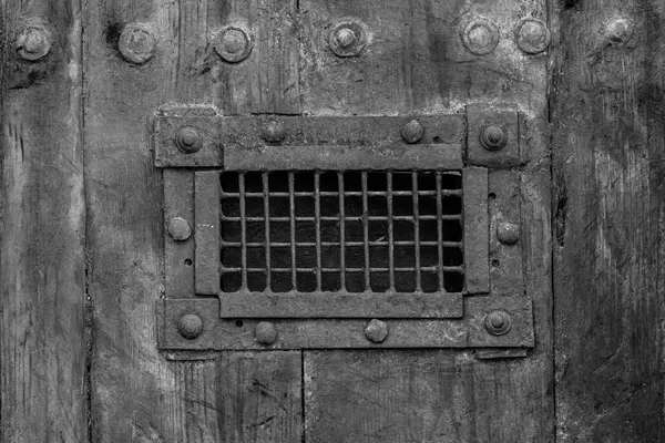 Portone di legno rinforzato da file di grosse borchie di ferro, con al centro uno sportello chiuso da una grata metallica dietro cui si vede solo buio, fotografia in bianco e nero
