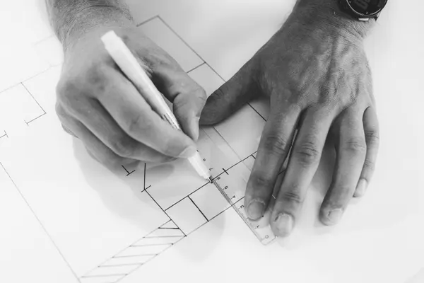 Due mani disegnano e misurano una pianta tecnica con penna e riga, in bianco e nero