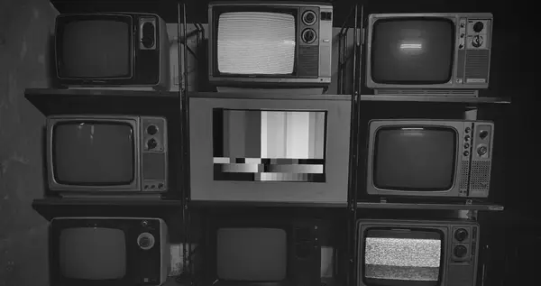 Parete di vecchi televisori impilati su scaffali, uno mostra il disturbo e uno una barra di calibrazione, fotografia in bianco e nero