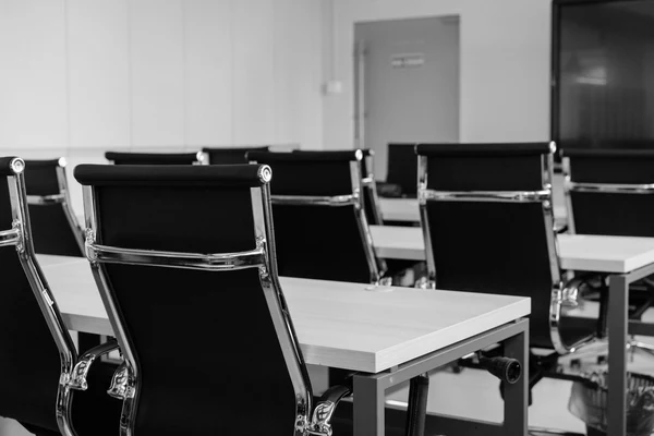 Fila di scrivanie con poltroncine da ufficio vuote in una sala illuminata, nessuno seduto, fotografia in bianco e nero