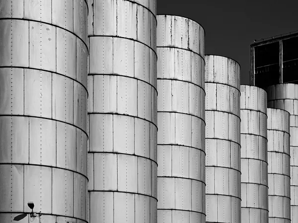 Fila di silos industriali cilindrici in metallo, ripresi da vicino in bianco e nero, con un edificio scuro sullo sfondo