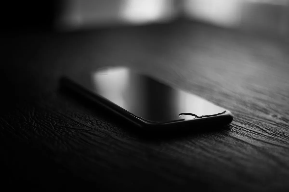Uno smartphone dallo schermo spento appoggiato su un piano di legno scuro, in bianco e nero