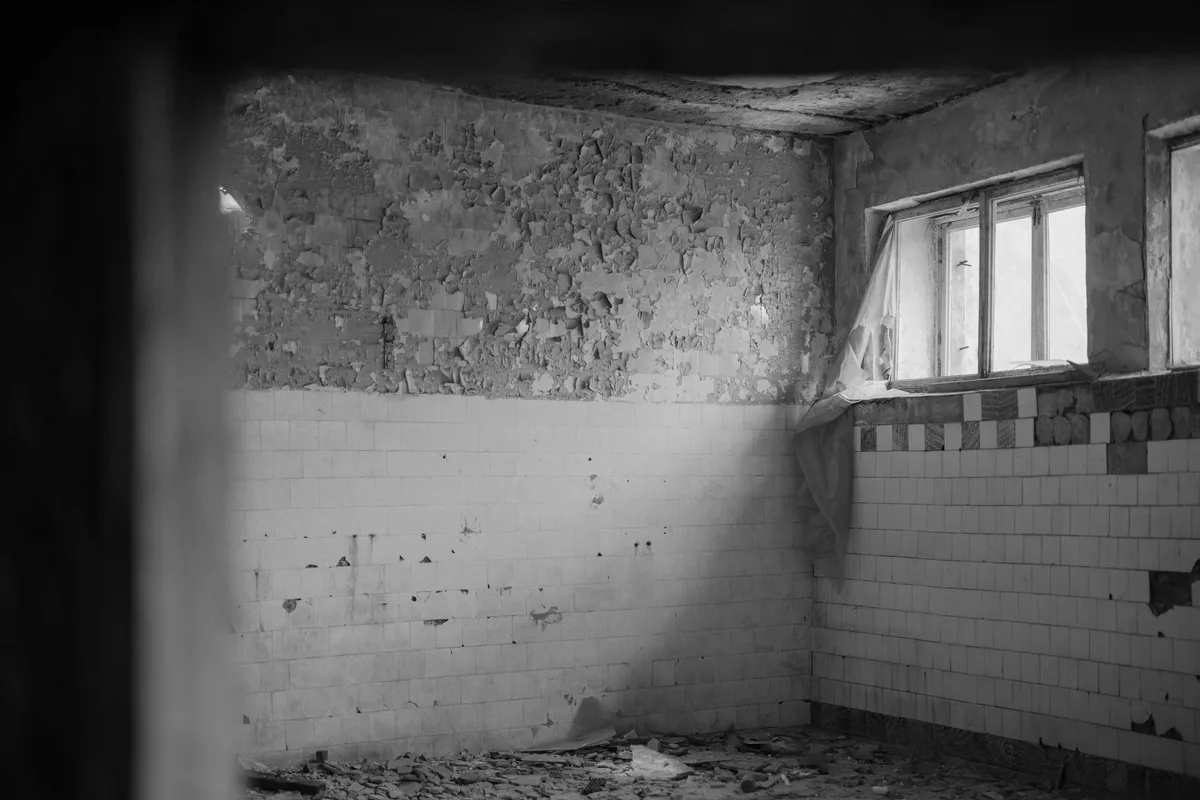 An empty, derelict room with tiled walls and fallen plaster, light from a window, black-and-white photograph with no people and no writing