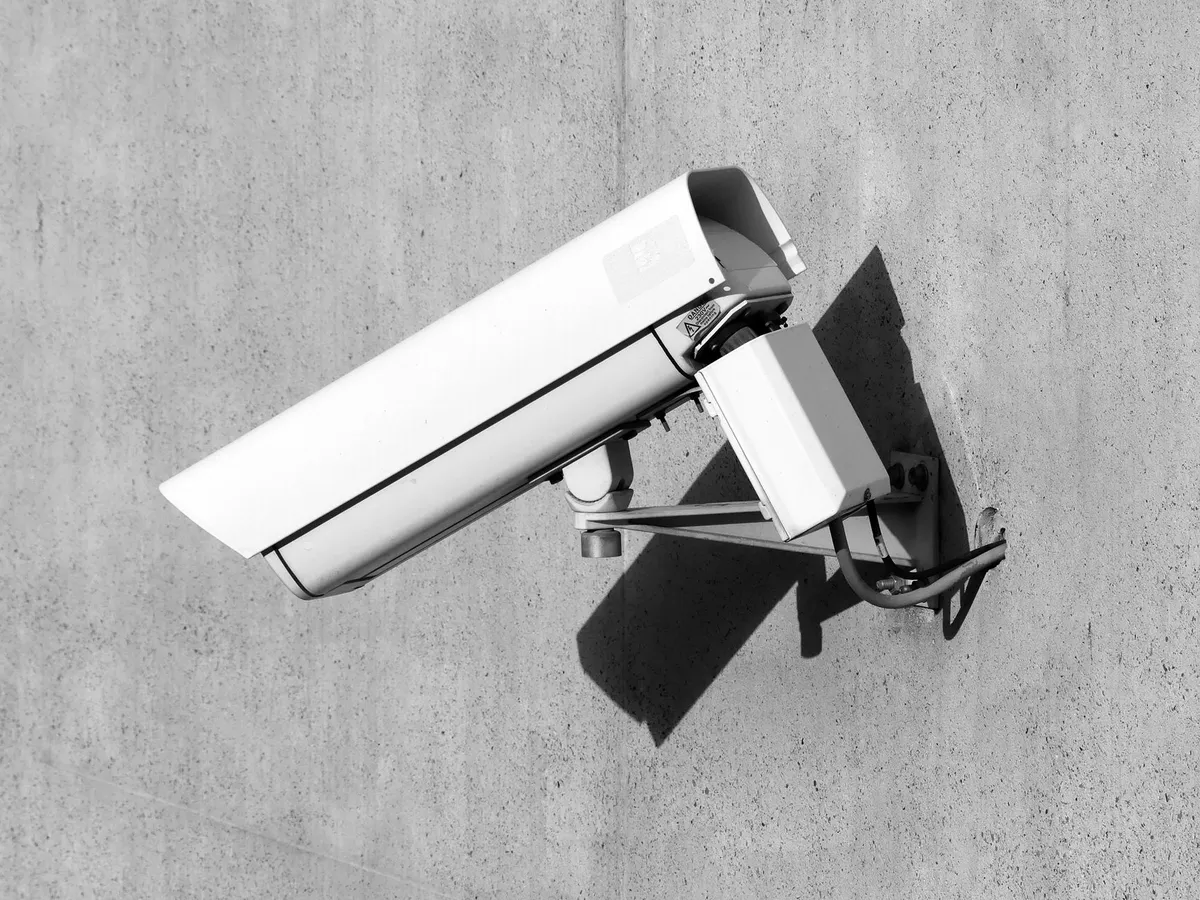 Telecamera di videosorveglianza bianca fissata a un muro di cemento, ripresa dal basso in controluce, in bianco e nero