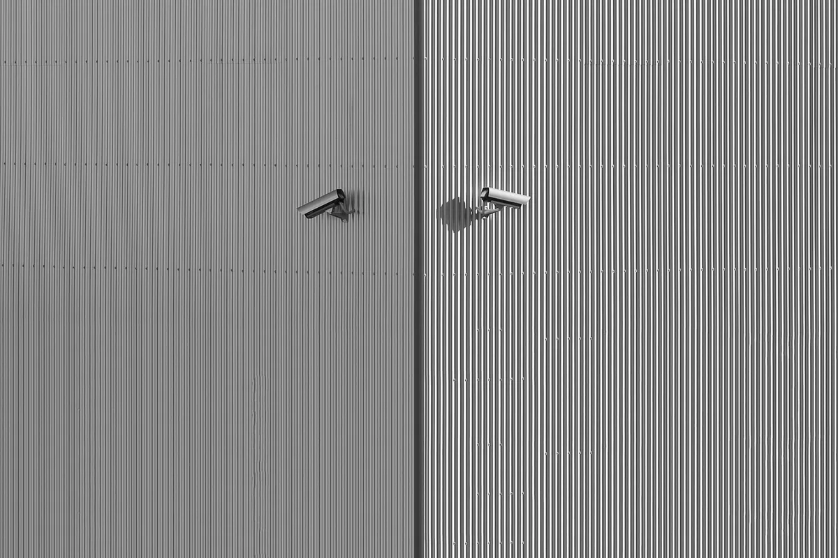 Two surveillance cameras fixed to a grey corrugated metal wall, in black and white