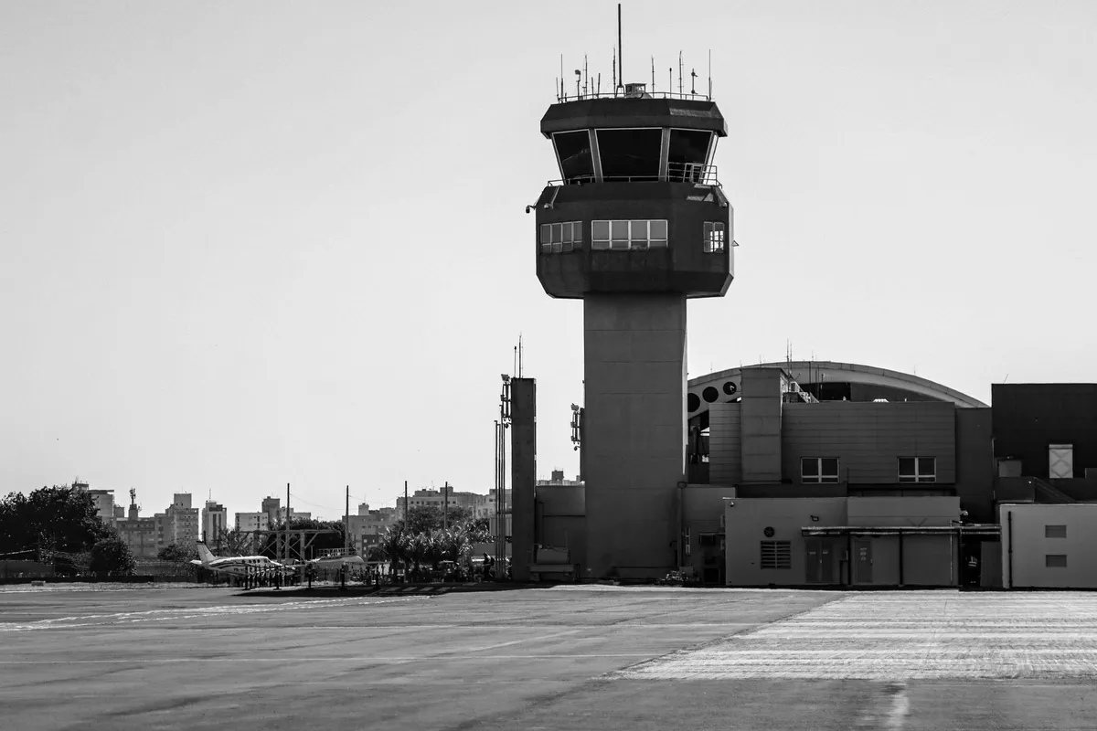 Torre di controllo del traffico aereo in bianco e nero, vista dalla pista di un aeroporto, con antenne sul tetto e un piccolo aereo in lontananza