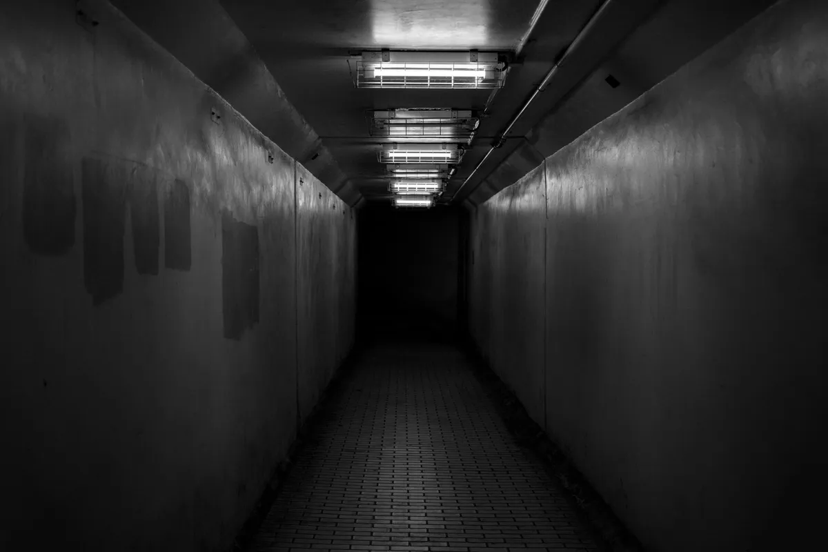 Corridoio stretto e buio, illuminato da una fila di tubi al neon sul soffitto, pavimento in mattonelle, in bianco e nero